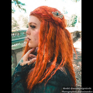 Model with vibrant orange hair styled with an ornate silver hair comb featuring a large black gemstone, standing in a sunlit garden with gothic-inspired attire.