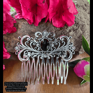 Intricately designed silver hair comb featuring a large black gemstone centerpiece and an ornate swirling motif, styled against a natural background with vibrant pink flowers and rustic stone.