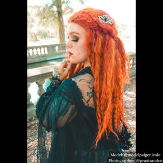 Model with vibrant orange hair styled with an ornate silver hair comb featuring a large black gemstone, standing in a sunlit garden with gothic-inspired attire.