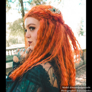 Model with vibrant orange hair styled with an ornate silver hair comb featuring a large black gemstone, standing in a sunlit garden with gothic-inspired attire.