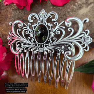 Intricately designed silver hair comb featuring a large black gemstone centerpiece and an ornate swirling motif, styled against a natural background with vibrant pink flowers and rustic stone.