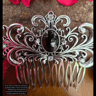 Intricately designed silver hair comb featuring a large black gemstone centerpiece and an ornate swirling motif, styled against a natural background with vibrant pink flowers and rustic stone.