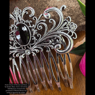 Intricately designed silver hair comb featuring a large black gemstone centerpiece and an ornate swirling motif, styled against a natural background with vibrant pink flowers and rustic stone.