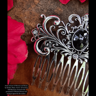 Intricately designed silver hair comb featuring a large black gemstone centerpiece and an ornate swirling motif, styled against a natural background with vibrant pink flowers and rustic stone.