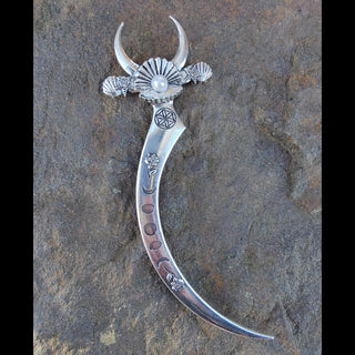 Ornate silver crescent-shaped hair dagger featuring a pearl centerpiece on the bed of a clam shell, moon phases, floral engravings, and mystical symbols, styled against a rustic stone surface with pink roses and white baby's breath flowers