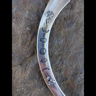 Ornate silver crescent-shaped hair dagger featuring a pearl centerpiece on the bed of a clam shell, moon phases, floral engravings, and mystical symbols, styled against a rustic stone surface with pink roses and white baby's breath flowers