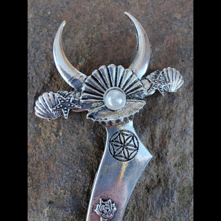 Ornate silver crescent-shaped hair dagger featuring a pearl centerpiece on the bed of a clam shell, moon phases, floral engravings, and mystical symbols, styled against a rustic stone surface with pink roses and white baby's breath flowers