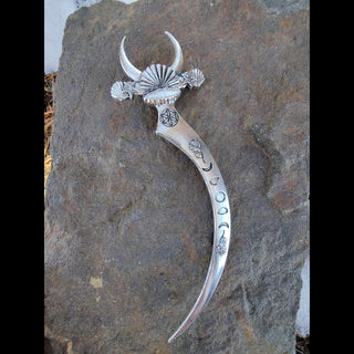 Back view of ornate silver crescent-shaped hair dagger featuring a pearl centerpiece on the bed of a clam shell, moon phases, floral engravings, and mystical symbols, styled against a rustic stone surface with pink roses and white baby's breath flowers