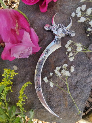 Ornate silver crescent-shaped hair dagger featuring a pearl centerpiece on the bed of a clam shell, moon phases, floral engravings, and mystical symbols, styled against a rustic stone surface with pink roses and white baby's breath flowers