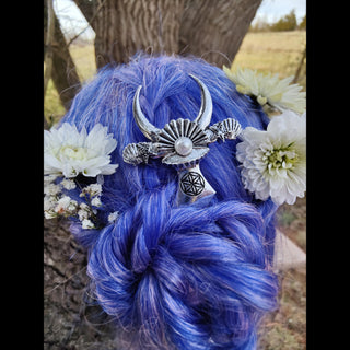 Silver crescent moon hair dagger with a pearl centerpiece and shell details, styled in vibrant braided purple hair adorned with white chrysanthemums and baby's breath, set against a tree trunk in a natural outdoor setting