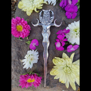 Full view of a silver hair stick featuring an owl perched between crescent moons, with a flowing, wavy design and engraved arrows, styled on a natural stone surface surrounded by colorful flowers