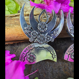 Close-up of the matching best friend pendant with a crescent moon engraved with "Merry We Part Friends," highlighting its floral accents and displayed with bright pink flowers on a natural wooden surface