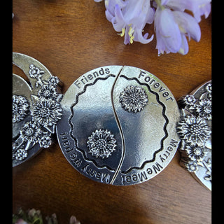 A detailed view of the two pendants joined together to form a yin-yang-inspired design, engraved with "Friends Forever" and  Merry We Meet, Merry We Part" and embellished with floral details, surrounded by lilac flower