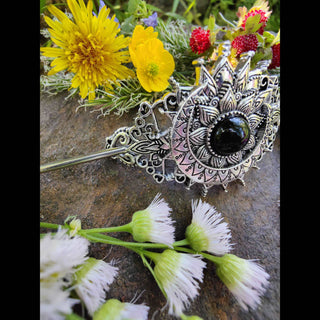 Close-up of a crescent moon hair stick with a detailed Victorian-style design, emphasizing the black gemstone centerpiece and ornate filigree patterns, set against a backdrop of wildflowers and freshly picked greenery on a textured stone