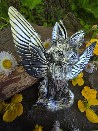 Angled view of the silver fox hair clip with spread wings, showcasing the detailed texture of the fox’s fur and wings, surrounded by vibrant yellow buttercups and white wildflowers on a stone surface with greenery in the background