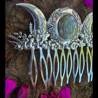 Side angle of a silver hair comb featuring a central labradorite stone and crescent moon accents, adorned with intricate flowers. The comb is surrounded by scattered magenta flower petals on a rock surface, emphasizing its celestial and botanical theme