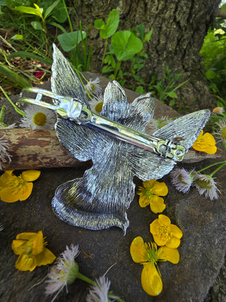 ack view of a silver winged fox-shaped hair clip, highlighting the intricate etched texture of the wings, displayed on a textured stone surface with scattered buttercup blossoms and delicate white wildflowers, set against a natural forest backdrop