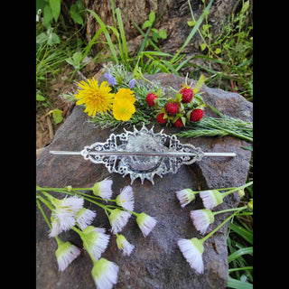 The back view of a crescent moon hair stick featuring Victorian-inspired filigree and a sturdy metal pin, displayed on a textured stone surface surrounded by vibrant dandelions, buttercups, wild strawberries, and delicate white wildflowers