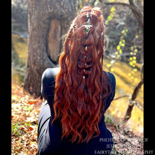 Back view of long, wavy red hair styled with intricate braids and adorned with a sword-shaped hair stick featuring a green gemstone and detailed filigree, set in a natural wooded area with sunlight filtering through the trees