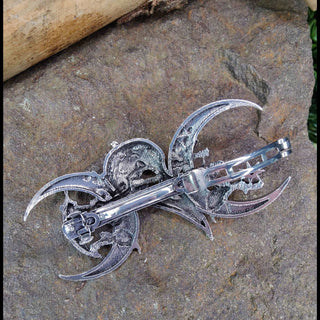 The backside of a silver crescent moon and raven hair barrette, displaying its intricate construction. The clip mechanism and delicate engraving details on the reverse side are visible, all set against a textured stone backdrop.