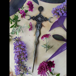  A close-up of the bronze finish Hekate-inspired hair dagger lying flat on a wooden surface adorned with purple and pink flowers, showcasing crescent moons, torches, and detailed engravings.