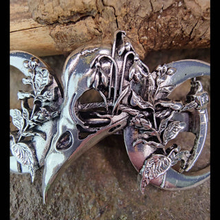 A close-up of a silver-tone crescent moon barrette adorned with a central raven skull. Surrounding the skull are finely etched leaves and flowers, highlighting the natural and mystical elements of the design. The barrette sits against a rough wooden surface.