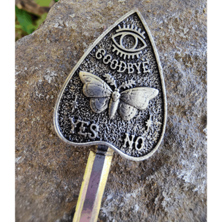 Close-up of a planchette-shaped hair stick, featuring intricate carvings of a moth with wings spread, an eye symbol above it, and the words "Goodbye," "Yes," and "No" engraved in textured metal against a stone background.