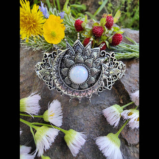 Crescent moon-shaped hair stick with a luminous opalescent stone surrounded by intricate Victorian-inspired designs, set against a natural backdrop of freshly picked dandelions, buttercups, wild strawberries, and soft white wildflowers