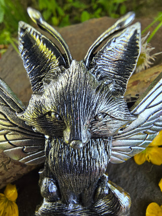 Close-up of the silver fox figure on the hair clip, emphasizing its finely etched fur texture, pointed ears, and serene expression, displayed against a natural backdrop with scattered wildflowers and greenery