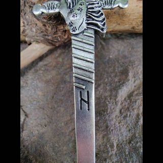 A close-up of the hair sword's blade shows horizontal engravings, symbolizing Hel's spiral, near the hilt and an "H" rune closer to the tip. The texture of the silver finish and the precise etching are clearly visible, with the sword resting on a weathered wood surface