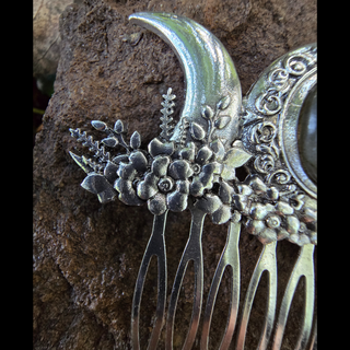 Detailed close-up of the left crescent moon on a silver hair comb, highlighting the floral engravings. The texture and craftsmanship of the flowers, along with the comb's metallic sheen, are accentuated against the natural rock backdrop