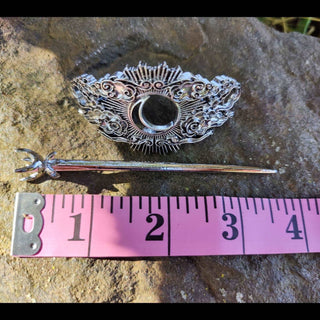 A silver hair cage featuring a crescent moon surrounded by intricate swirling clouds and sunburst rays, resting on a stone with a pink measuring tape for scale