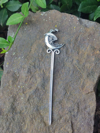  A silver-toned hair stick featuring a crescent moon design with a curled dragon resting on its surface, set on a textured stone background surrounded by greenery.