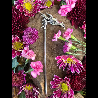 Silver dragon skull hair scythe placed on a rocky surface surrounded by vibrant pink and purple flowers