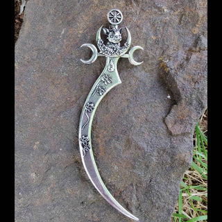 A full-length view of the silver curved hair dagger inspired by the Goddess Freya, displayed on a textured stone surface. The handle is engraved with intricate floral patterns and gracefully curves into a sharp dagger-like point. The boar motif, framed by moon-shaped daggers, reflects Freya's connection to nature, power, and mysticism, making it a striking, symbolic piece.