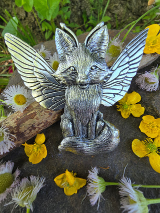 Full view of the silver fox hair clip with outstretched wings, seated on a stone surface surrounded by freshly picked yellow buttercups and soft white blossoms, set against a lush, green forest backdrop