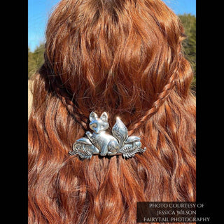 Silver fox-shaped hair barrette securing braided red hair, with the intricate design of the fox and its engraved tail illuminated by natural sunlight, outdoors against a blurred natural backdrop