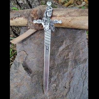 The full-length hair sword is displayed on a textured rock, its blade and handle fully visible. Hel’s dualistic depiction and the engraved lines on the blade add character to the piece, with natural wood and greenery creating an earthy background.