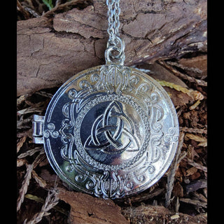 The silver Celtic locket displayed on a bed of brown bark, with its intricate triquetra design and surrounding ornate engravings shining under natural light. The fine details of the engravings stand out against the textured bark surface, creating a harmonious contrast.