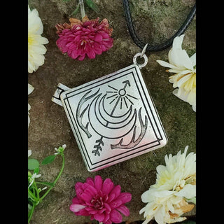 A single square locket pendant resting on moss and surrounded by vibrant flowers in shades of white, cream, and pink. The locket is closed and features a crescent moon design with antler-like details and celestial rays.