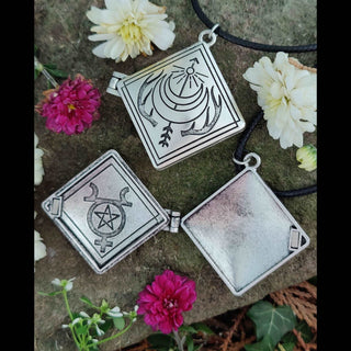 Two diamond-shaped locket pendants displayed on a mossy surface. One locket is open, revealing intricate celestial and pentacle engravings, while the other displays an engraved crescent moon with antler-like details on its front. The arrangement is surrounded by delicate white, cream, and pink flowers.