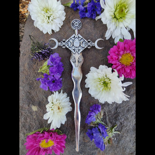 The Hekate-inspired hair dagger displayed in its entirety on a textured stone surface, surrounded by a vibrant arrangement of fresh flowers in shades of white, magenta, purple, and soft yellow. The dagger features a flowing, serpentine blade with a metallic sheen that catches the light, leading to a detailed hilt adorned with engraved torches and crescent moons. The geometric centerpiece of the hilt showcases a triple moon symbol, representing Hekate's divine connection to the phases of the moon, while deli