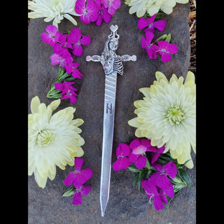 The hair sword is surrounded by vibrant flowers in shades of white, cream, and magenta, enhancing its ethereal quality. The detailed design of the blade, the "H" rune, and the goddess Hel’s symbolic depiction stand out against the contrasting natural elements.