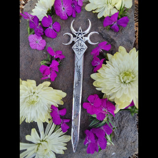 The Nyx-inspired hair dagger surrounded by bright flowers on a stone surface. The celestial design stands out against the vivid purple, yellow, and white petals, emphasizing its contrast of dark elegance and natural beauty.