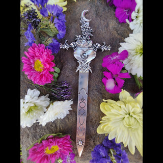 The Persephone-inspired hair dagger in its full glory, placed against a rocky backdrop surrounded by vibrant flowers in shades of pink, purple, and white. The blade’s geometric engravings lead up to the ornate floral crest with a crescent moon and heart centerpiece, with a sleeping faun nested amonst the flowers. creating a striking visual representation of rebirth and duality.