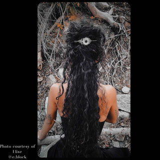 Artistic photograph of long, curly black hair styled with a crescent moon hair stick featuring a black gemstone centerpiece and ornate filigree details, worn by a model seated in a forested area with exposed roots and rugged terrain