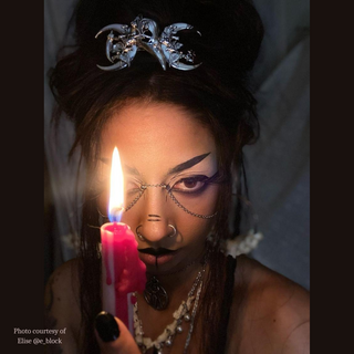 The same model gazes directly into the camera while holding the candle, with the raven skull barrette adding an elegant, mystical touch to their hair. The dark, atmospheric lighting enhances the overall aesthetic.