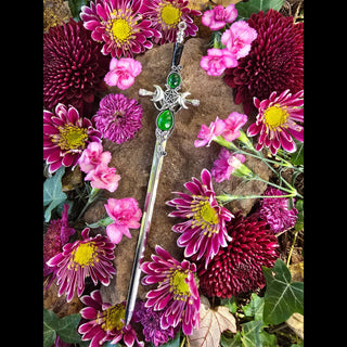 Decorative sword-shaped hair stick with crescent moons, green gemstones, and pentacle designs, displayed on a textured stone with a vivid arrangement of magenta and pink chrysanthemums, carnations, and ivy leaves.