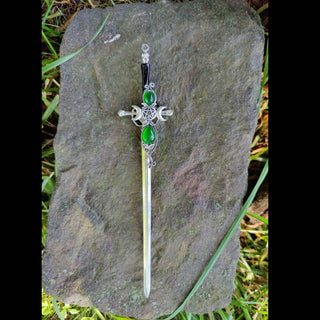 Decorative sword-shaped hair stick with crescent moons, green gemstones, and pentacle designs, displayed on a textured stone with a vivid arrangement of magenta and pink chrysanthemums, carnations, and ivy leaves.