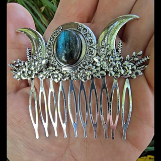 A single silver hair comb resting in the palm of a hand to showcase its size and craftsmanship. The design features a triple moon sigil with a central labradorite stone, surrounded by ornate floral details, blending mysticism with nature-inspired elements